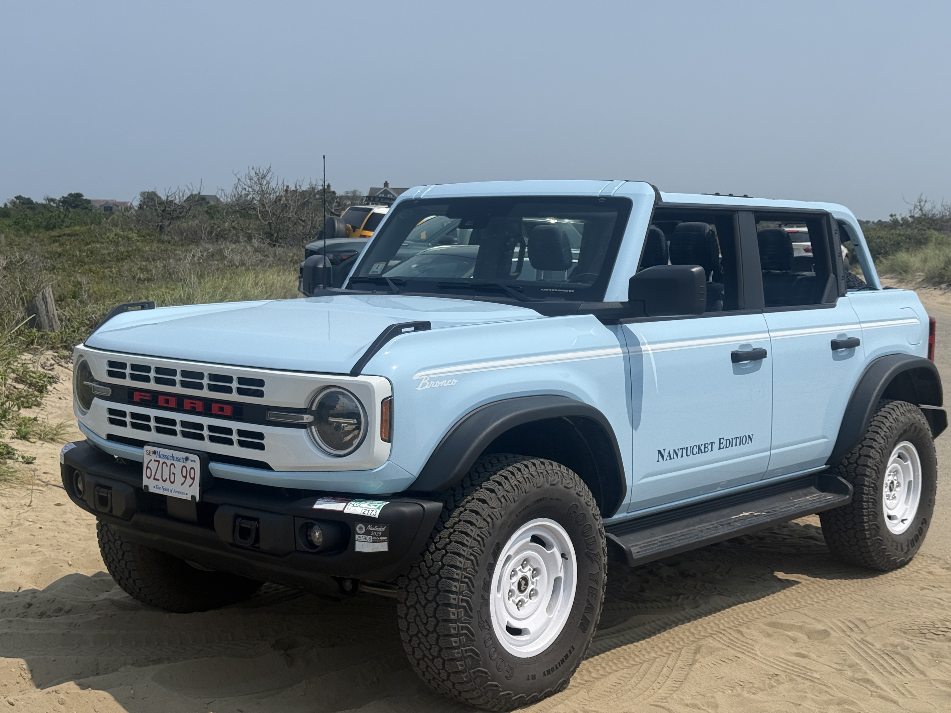 Light blue Ford Bronco Nantucket Edition with the top off, parked on the sand beside dune grass
