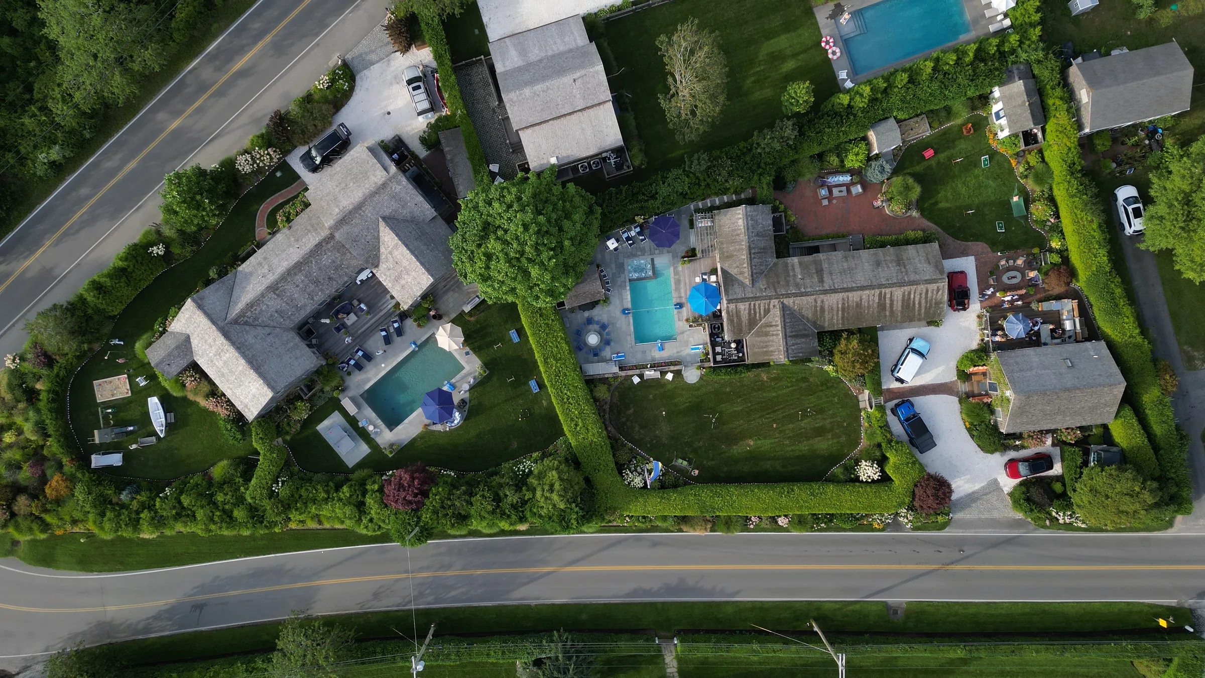 Top-down aerial of the compound: the shingled houses on one set of hedged, garden-lined grounds, each with its own pool and terrace, bounded by two lanes.