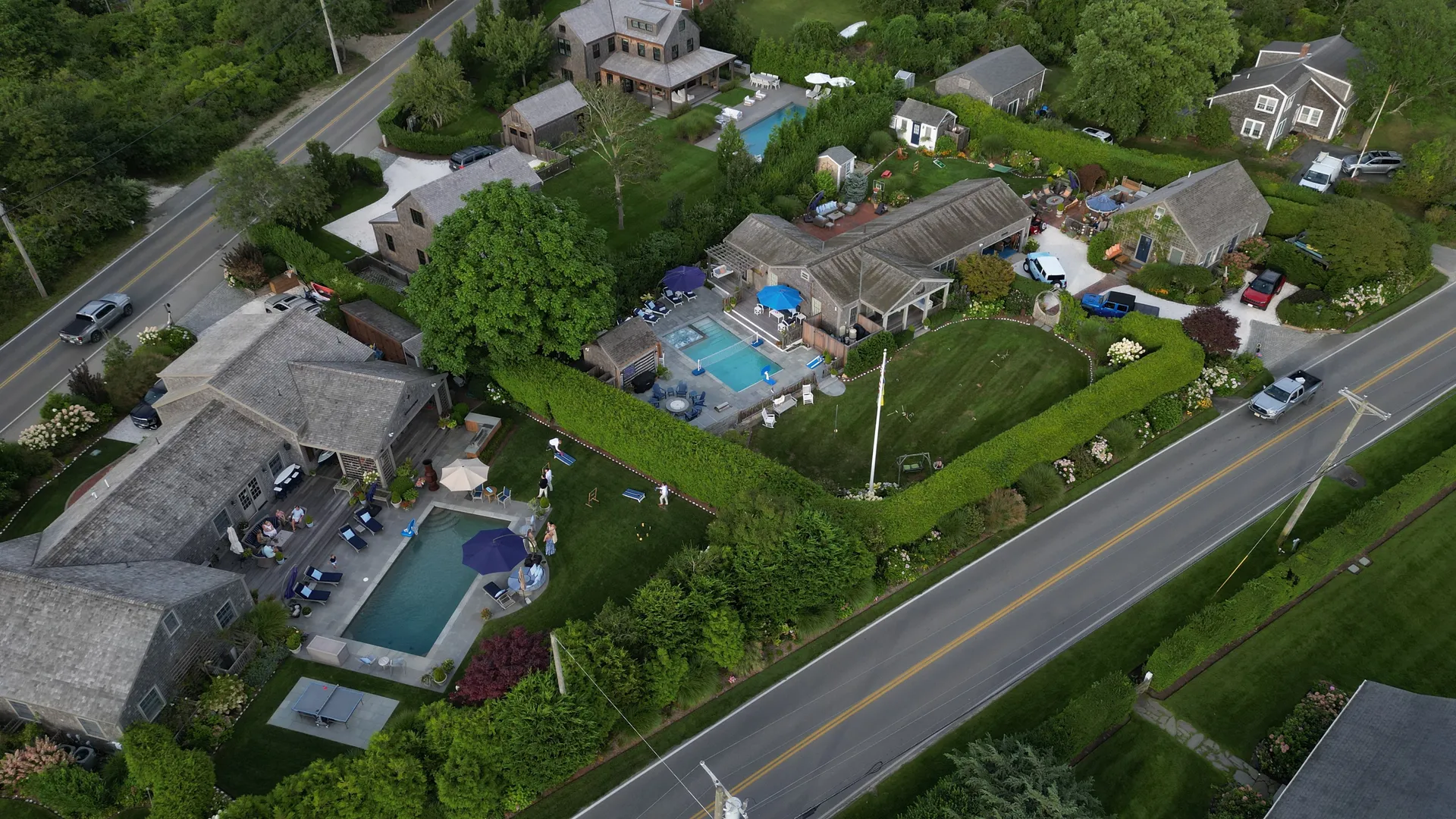 Aerial view of the whole compound — the three cedar-shingled houses set among clipped hedges and gardens, each with its own pool and bluestone terrace, linked by lawns and gravel drives.